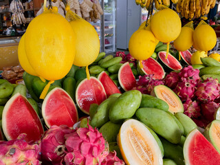 A colorful and bountiful display of fresh tropical fruits, including bright yellow melons, red watermelons, and pink dragon fruits, all arranged on a stall at a traditional market.

