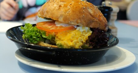 Burger features scrambled egg on table against visitor eating in restaurant. Ingredients sandwiched between toasted sesame seed bun haves exuding nutty aroma