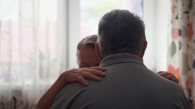 A sad elderly couple sits hugging near the window of a house