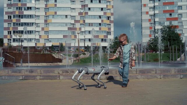 Young boy in sunglasses approaching and petting a futuristic four-legged robot dog in a city park