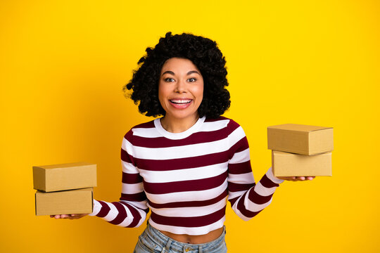 Fototapeta Smiling young woman holding cardboard boxes in both hands against a yellow background wearing a striped pullover
