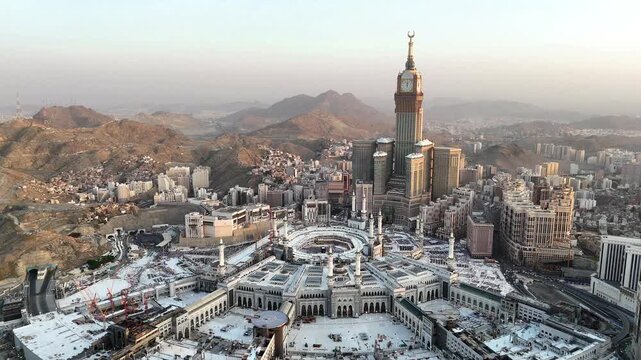 drone footage of Mecca, Saudi Arabia, filmed at sunset. The video captures the vast urban landscape with residential buildings, modern towers, and surrounding mountains bathed in golden hour light