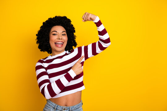 Fototapeta Cheerful young woman with curly hair pointing at bicep in striped outfit against a bright yellow background