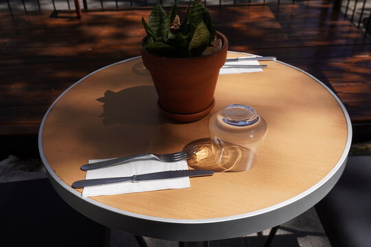 Round outdoor caf&eacute; table set with cutlery on napkins, an upside-down glass, and a potted plant as decoration. Sunlight creates soft shadows, giving the scene a cozy and inviting atmosphere