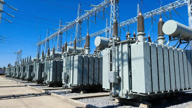Row of high-voltage electrical transformers at a power substation. Industrial infrastructure with metal structures, cables, and equipment under a clear blue sky.