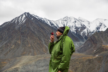 A man, European, 50 years old, stands with a walkie-talkie in his hands against a backdrop of mountain peaks. He's talking on the radio.