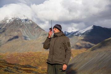 A man, European, 50 years old, stands with a walkie-talkie in his hands against a backdrop of mountain peaks. He's talking on the radio.