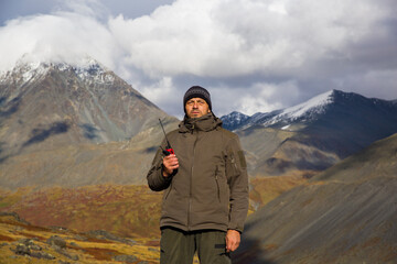 A man, European, 50 years old, stands with a walkie-talkie in his hands against the backdrop of mountain peaks.