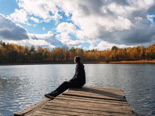 A man sits on a wooden bridge by the lake and admires the autumn landscape.