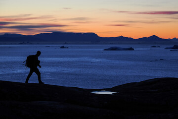 Silhouette of a lone hiker with a backpack walking on rocky shore at twilight with icebergs and mountains in Disko Bay, Greenland