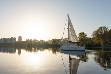White sailing yacht with tall mast and raised sails on still river water near forested autumn shore. Peaceful leisure activity and travel concept in nature with golden fall trees.
