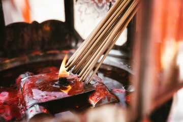 A close-up shot of several incense sticks being held over a flame in a traditional metal holder, covered in molten red wax or oil, during a Buddhist prayer or ritual.