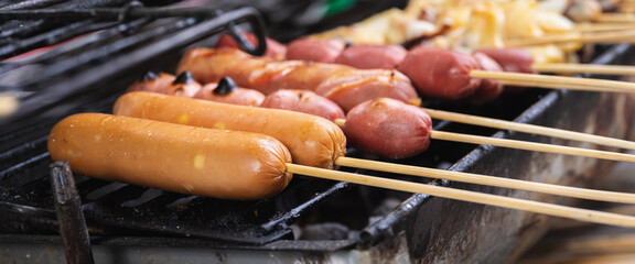 A close-up shot of various sausages and hot dogs on wooden skewers grilling over hot coals on a black barbecue grill. This image represents street food and outdoor cooking.