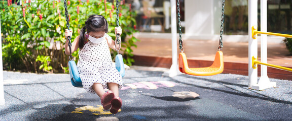A candid of young Asian child girl wearing a white face mask is sitting on a swing at an outdoor playground. Childhood during a global pandemic. Sunny park setting with lush green foliage.