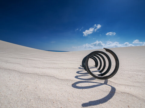 Rusty metal spring on white sand casting spiral shadow in bright desert under blue sky