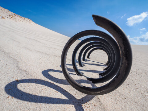 Rusty metal spring on white sand casting spiral shadow in bright desert under blue sky