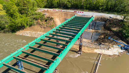 Aerial view of a partially constructed green steel bridge over a river, surrounded by lush greenery. Heavy machinery is visible nearby, indicating ongoing construction and development.