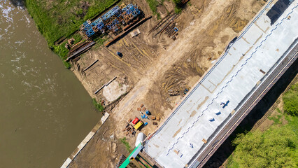Aerial view of construction site alongside a river, featuring equipment and building materials. The area shows soil and machinery, indicating active construction.