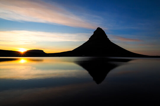 Pointed mountain silhouette reflecting in still water during vivid Icelandic sunset under colorful sky. Kirkufjell Iceland