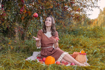 Portrait of a woman tossing an apple while sitting on a picnic blanket in autumn grass, surrounded by rowan berries, apples and a melon