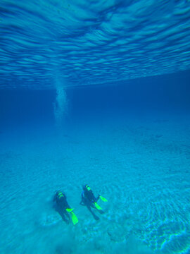 Two scuba divers underwater on a sandy seabed with clear blue water and dramatic light reflections above