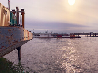 Weathered barge with a small cabin, moored on calm harbor water. In the background, cranes, ships and port infrastructure fade into hazy golden-hour sky with soft sun flare. Maritime/industrial scene