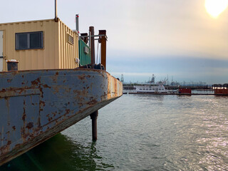 Weathered barge with a small cabin, moored on calm harbor water. In the background, cranes, ships and port infrastructure fade into hazy golden-hour sky with soft sun flare. Maritime/industrial scene