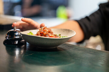 Close-up of a poke bowl with diced raw fish, sesame seeds, and herbs on a dark green counter. A hand reaches for a service bell, suggesting a restaurant or food service setting