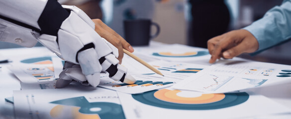 Robotic hand collaborates with human in analyzing various charts and graphs on papers, highlighting the integration of technology in modern business practices.Noogenesis