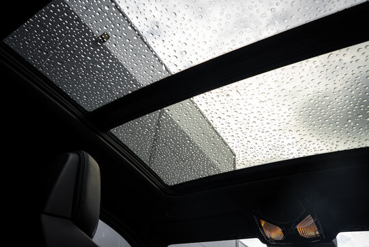 Interior view of a BMW X4 focusing on its panoramic sunroof covered in rain droplets, with a cloudy sky and modern building visible in the background.
