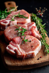 Fototapeta premium Fresh pork chops prepared for cooking on a wooden cutting board with herbs set against a dark background