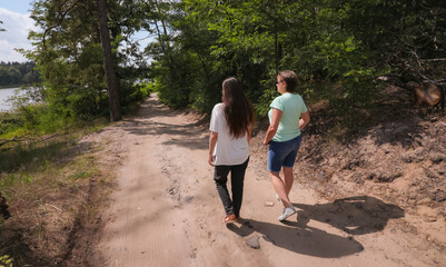 Two women walking along a sandy forest path near a river on a sunny day, enjoying nature and conversation. Friendship, leisure, and outdoor lifestyle.
