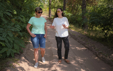 Obraz premium Two women walking on a sandy forest path, wearing sunglasses and casual clothes, talking and enjoying a sunny day outdoors. Friendship, leisure, and lifestyle.
