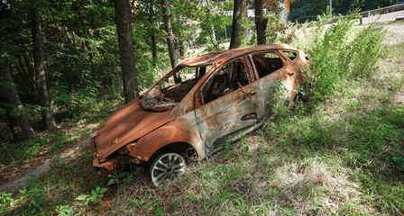 Abandoned burnt-out car wreck left in the forest near a road, covered in rust and surrounded by...