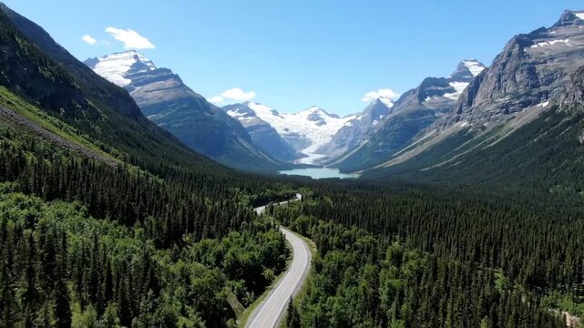 Aerial view of a scenic road winding through a lush green forest in glacier national park, montana, with snowcapped mountains in the background on a sunny day