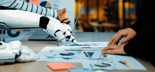 A futuristic robot collaborates with a human in an office setting, analyzing charts and data. Innovation and artificial intelligence enhance business productivity and teamwork.Noogenesis