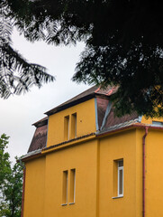 Close-up of a striking, bright yellow historic building's facade on a cloudy day, featuring unique double vertical windows and a weathered, dark red mansard roof peaking out from behind dark green tre