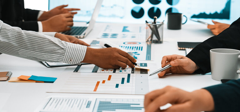 A diverse business team engages in active collaboration at a modern office setting. They analyze charts and graphs while discussing strategies over laptops and documents. SACTR