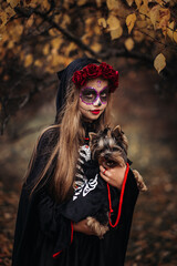 A young woman in a Day of the Dead costume: purple and violet skull makeup, red roses on a wreath, and a black cloak. She held a small dog against the backdrop of an autumn park with fallen leaves.