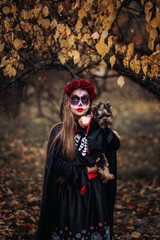 A young woman in a Day of the Dead costume: purple and violet skull makeup, red roses on a wreath, and a black cloak. She held a small dog against the backdrop of an autumn park with fallen leaves.