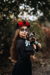 A young woman in a Day of the Dead costume: purple and violet skull makeup, red roses on a wreath, and a black cloak. She held a small dog against the backdrop of an autumn park with fallen leaves.