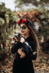 A young woman in a Day of the Dead costume: purple and violet skull makeup, red roses on a wreath, and a black cloak. She held a small dog against the backdrop of an autumn park with fallen leaves.