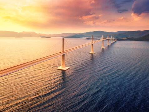 Aerial view of modern Peljesac Bridge and blue sea at colorful sunset in summer. Croatia. Top drone view of bridge during golden hour, mountains, reflection in water and orange sky. Architecture