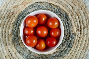 Cherry tomatoes fill a white bowl placed on a woven mat. Their vibrant red color contrasts with the neutral tones of the mat, highlighting their freshness and natural appeal.
