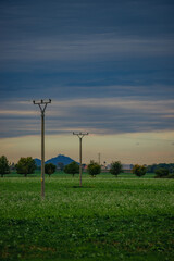 Obraz premium Minimalist rural landscape with power lines crossing a vast green field and a prominent hill with a fortress silhouette on the horizon under a soft blue and orange sunset sky.