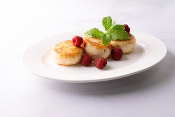 Cottage cheese pancakes with honey, raspberry and mint leaves on white ceramic plate on white marble table close up. Healthy homemade dessert.