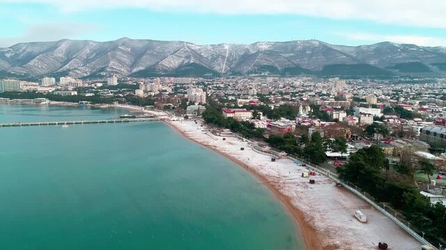 Central beach of Gelendzhik in snow. Aerial view. Zia at resort. Caucasus Mountains in snow. In Gelendzhik Bay you can see pier of sea station. People walking on snowy beach.