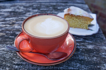 A large cup of latte with frothy milk in a pink ceramic cup and saucer with a lemon cake with icing on the top placed on a rustic wooden table.