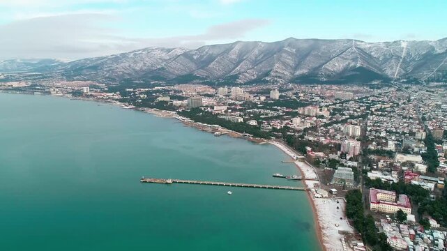 Central beach of Gelendzhik in snow. Aerial view. Zia at resort. Caucasus Mountains in snow. In Gelendzhik Bay you can see pier of sea station. People walking on snowy beach.