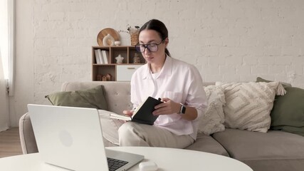 Woman in glasses sits on sofa with laptop and notebook, writes with pen and looks focused. Cozy home interior with pillows, shelf decor, books, vases, wicker basket, daylight and relaxed atmosphere
